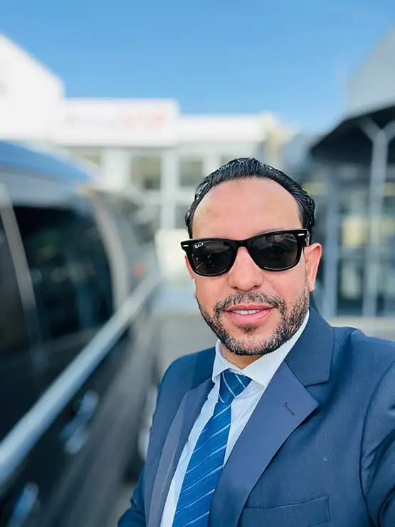 Chauffeur en costume bleu et lunettes de soleil posant à côté d’un véhicule.