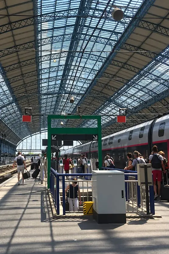 Voyageurs sur un quai de gare sous une grande verrière, avec un train à quai.